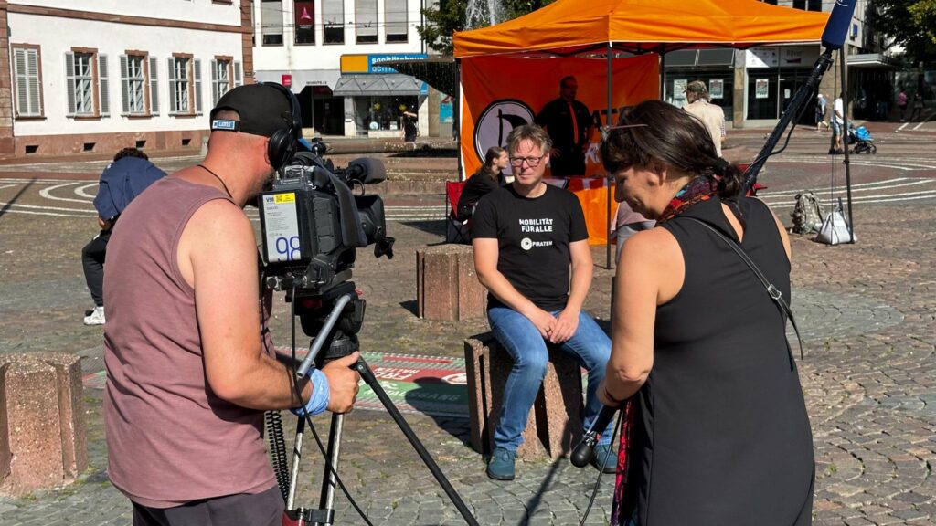 ein Kameramann filmt Sebastian vor einem Pavillon der Piratenpartei